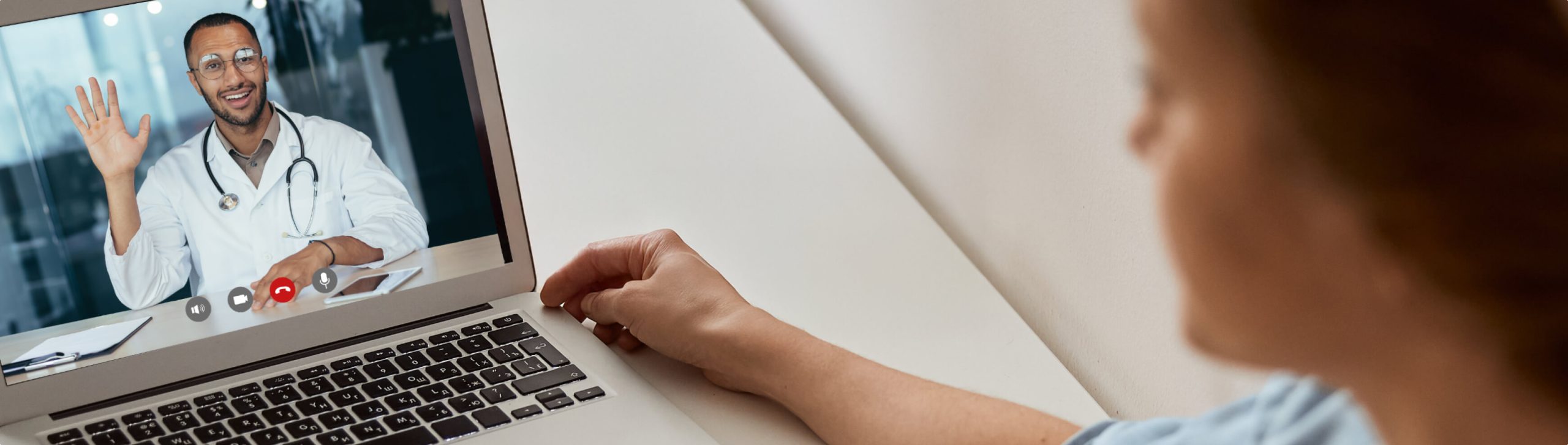 A person has an online doctor visit on a laptop. The doctor, wearing a white coat, is smiling and waving on the screen.