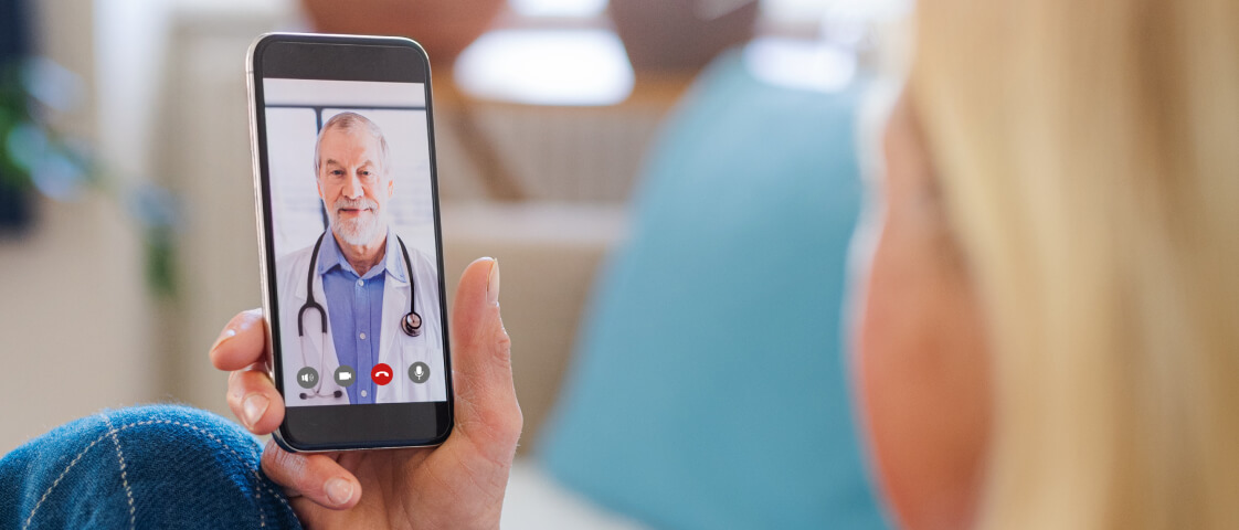 A person holds a smartphone during an online doctor visit, video calling a male doctor dressed in a white coat and stethoscope.