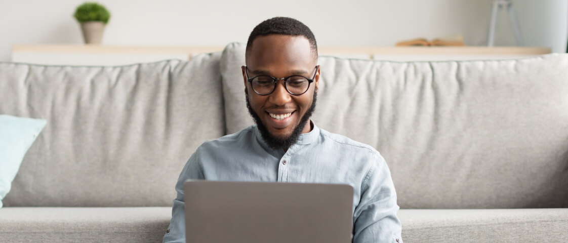 A man sits on a sofa smiling while having an online doctor visit on his laptop, with a plant and a book on a shelf in the background.