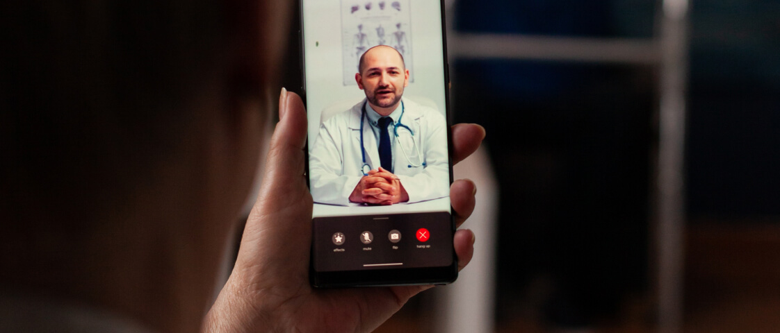 A person holds a smartphone showing a video call with a male doctor in a white coat and tie speaking, highlighting the convenience of an online doctor visit with telemedicine app icons visible on the screen.