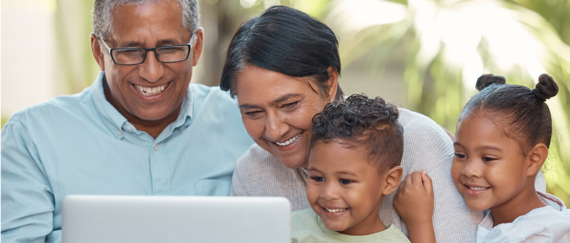 Two adults and two children sit close together, smiling and looking at a laptop screen in a bright indoor setting, connecting with Whole Person Virtual Care telehealth doctors.