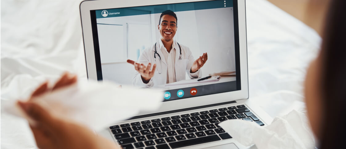 A person has a virtual consultation with a Whole Person Virtual Care telehealth doctor via video call on a laptop, holding tissues while the caring doctor gestures and smiles on the screen.