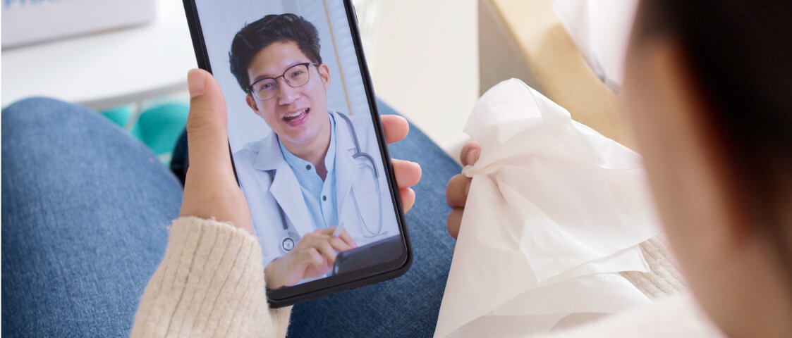 A person holds a phone and participates in a video call with a telehealth doctor wearing a white coat and stethoscope, while holding a tissue in their other hand—embracing the convenience of Whole Person Virtual Care.